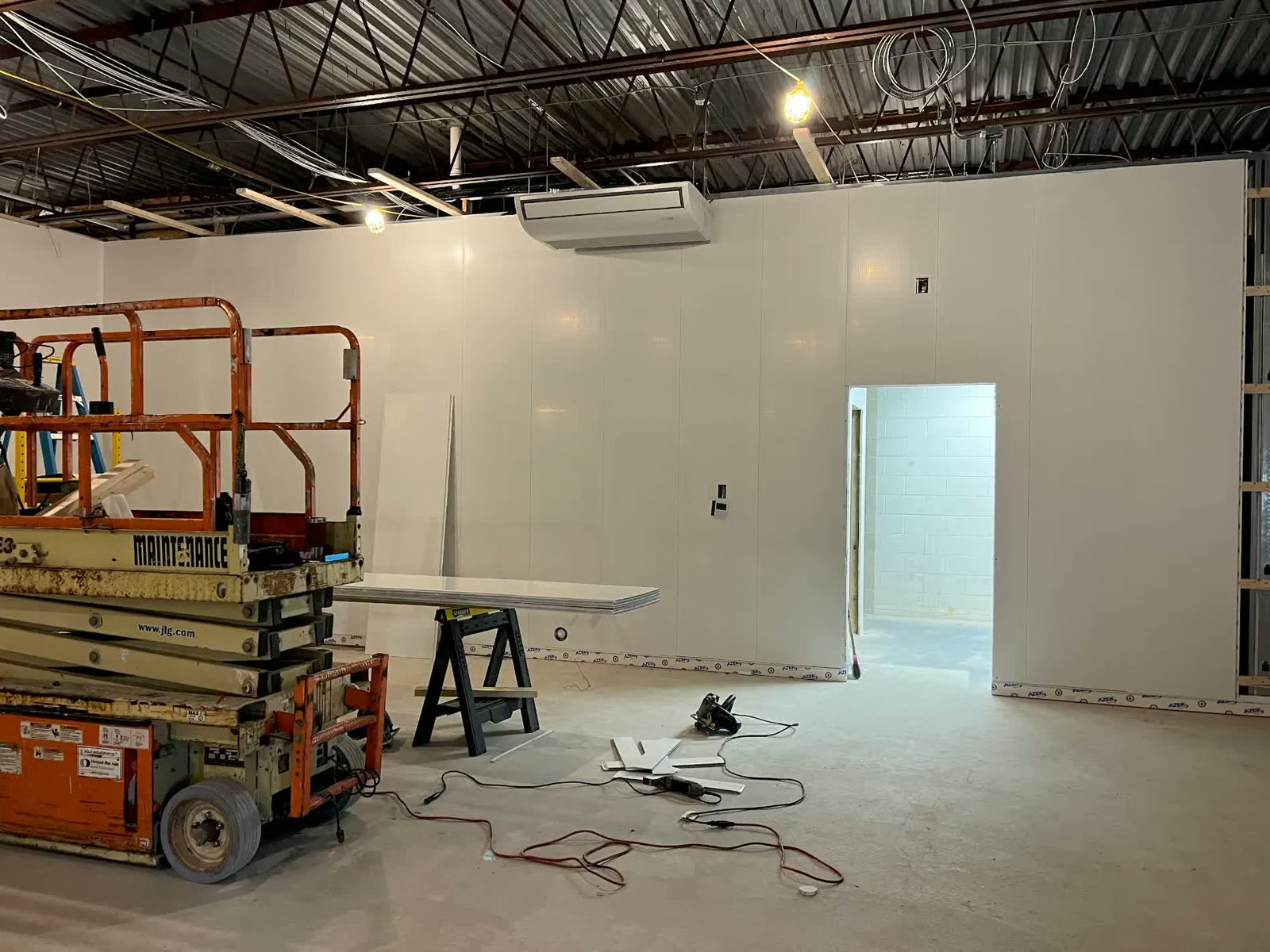 Commercial buildout in progress with new white wall panels, exposed ceiling joists, and a scissor lift